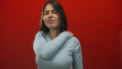 Woman experiencing neck pain with a pained expression, standing against an isolated red background,...