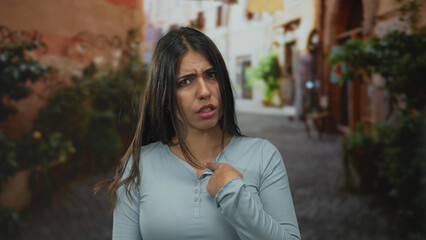 Young woman makes finger pointing gesture to herself on a narrow street in soft daylight; rejection.