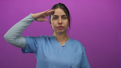 Young hispanic woman in blue scrubs saluting confidently against an isolated pink background,...