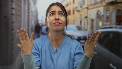 Woman in blue scrubs outdoors on street expressing frustration with blurred urban background and...