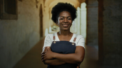Confident woman with arms crossed wearing apron stands indoors at an old university with arched corridors, conveying a sense of pride and assurance.