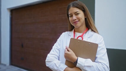 Young female doctor stands outdoors in an urban setting holding a clipboard, smiling with...