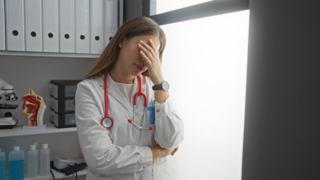 Woman doctor stressed in hospital room with stethoscope and files showing frustration in clinical environment during daytime.