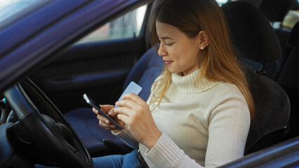 Woman sitting in car holding smartphone and credit card, smiling while shopping online against...