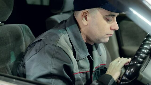Professional mechanic resting inside vehicle, scrolling smartphone screen during work pause at automotive service center, wearing workshop uniform