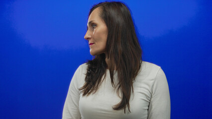 Woman with long hair wearing white shirt standing against vibrant blue background wall, gazing...
