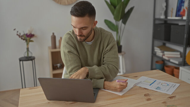 Young man working on laptop in a modern living room with charts on the table showcasing a productive home office environment. - Powered by Adobe