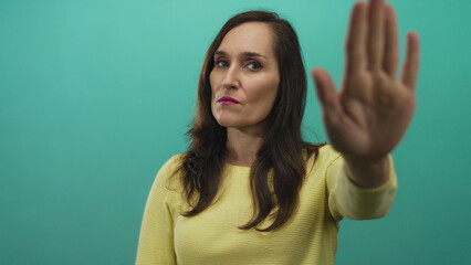 Woman with eyes closed extends hand in stop gesture against green background wearing yellow sweater...