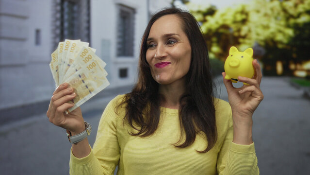 Woman holding swedish krona banknotes and a piggy bank outdoors in a street setting.