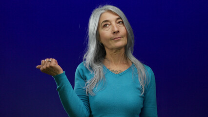 Fototapeta premium Senior woman with grey hair pointing sideways on isolated blue background wearing turquoise sweater expressing curious gesture