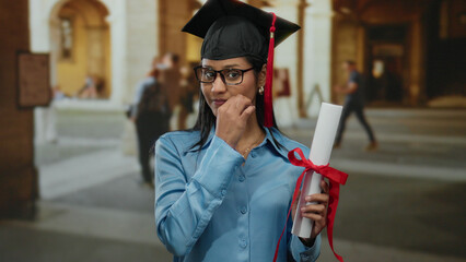 Young woman in blue shirt holding diploma and adjusting glasses in an old university indoor setting