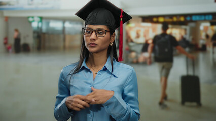 Young woman in graduation cap and glasses standing in an airport terminal with blurred travelers in...