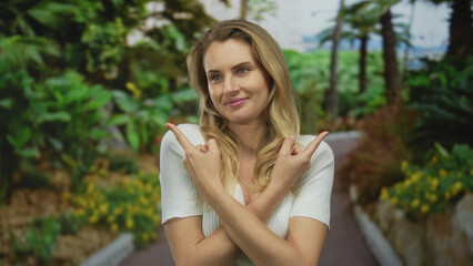 Young blonde woman pointing playfully in a lush green outdoor park setting with a cheerful expression amidst vibrant foliage and sunshine.