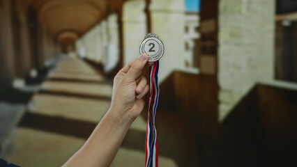 Hand holding silver medal indoors at historic university corridor showcasing award achievement in...