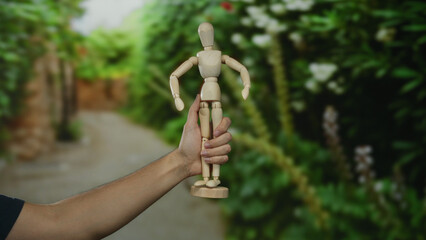 Man holding wooden mannequin in park background surrounded by greenery and flowers, showcasing concept of creativity and art outdoors.
