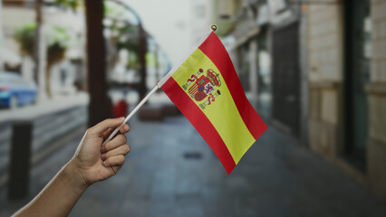 Hand holding a spanish flag in an outdoor town setting with a blurred street scene in the...