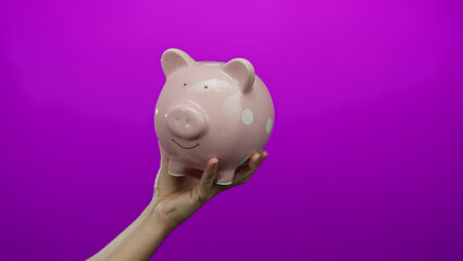 Caucasian hand holds a smiling pink piggy bank against a vivid pink wall, symbolizing savings and...