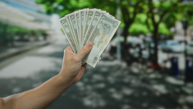 Caucasian man holding dirham banknotes in hand outdoors on a city street illustrating spending or budgeting.