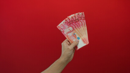 Caucasian male hand holding filipino pesos against vivid red wall offers a striking contrast and...