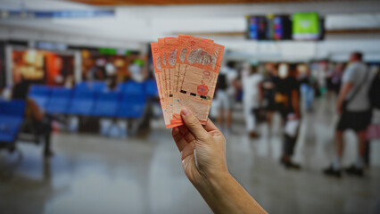 Hand holding malaysian ringgits in an airport terminal, with travelers moving in the background,...