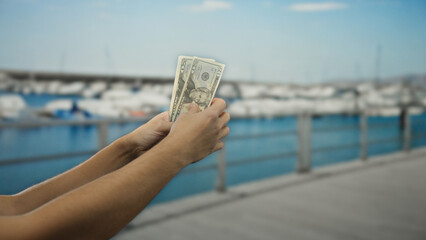 Man holding american dollars by the seaside with boats visible in the background, suggesting travel...