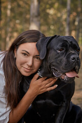 Obraz premium Young woman with cane corso dog sitting together in forest. Happy pet and smiling woman enjoying nature. Summer forest background, companionship, love for animals, responsible dog ownership.