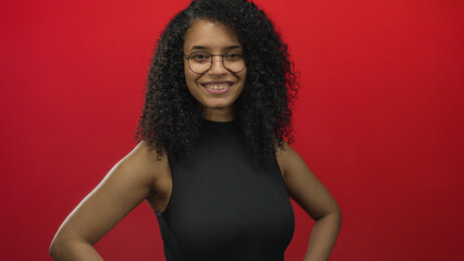 Woman smiling with curly hair and glasses in front of a vibrant red background, embodying...