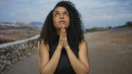 Woman praying outdoors with curly hair, wearing glasses, and a black shirt against a scenic nature...