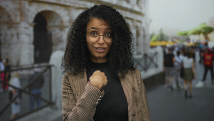 Woman with curly hair and glasses pointing finger to self on street in front of building facade; rejection.