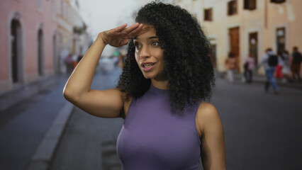 Young woman with hand shading eyes looking into distance down urban street lined with buildings;...