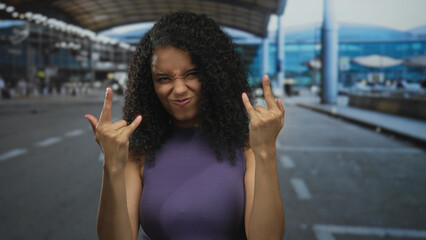 Woman making rock gesture while smiling outdoors at busy airport terminal with modern architecture in background, showcasing energy and excitement.