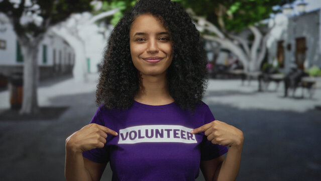 Woman with curly hair wearing volunteer shirt giving thumbs up on a street backdrop outdoors showcasing positivity and community spirit