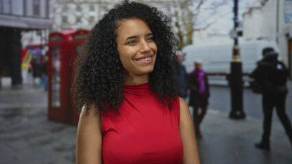 Woman smiling on london street with red telephone booth in background, wearing red top, enjoying...