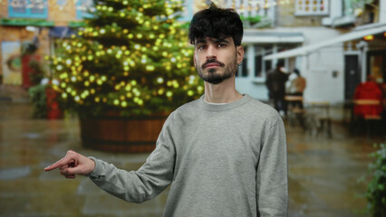 Young man in an urban street pointing near a brightly lit christmas tree outdoors in a city setting.