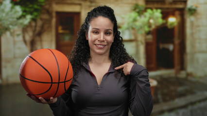 Woman holding basketball pointing at herself in lush urban street setting, young hispanic smiling...