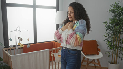 Woman in bedroom holding baby clothes beside crib with plants and chair in a bright morning light setting showcasing calm domestic indoor scene