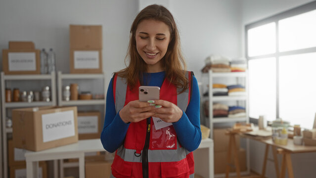 Young woman volunteer using phone in charity donations center room with boxes in the background showcasing a community support environment indoors.