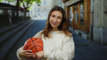 Woman holding basketball while smiling in urban street during daytime with trees in background.