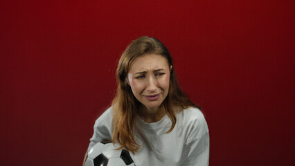 Young woman holding soccer ball appears sad against isolated red background wall, wearing white...