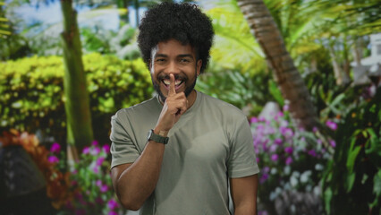 African american man in park outdoor making silence gesture surrounded by lush green trees and vibrant flowers implying tranquility and serenity in nature.