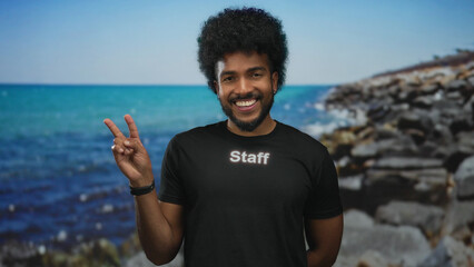 Smiling man in staff uniform flashing peace sign on a picturesque seaside outdoor setting with rocks and vibrant sea background.