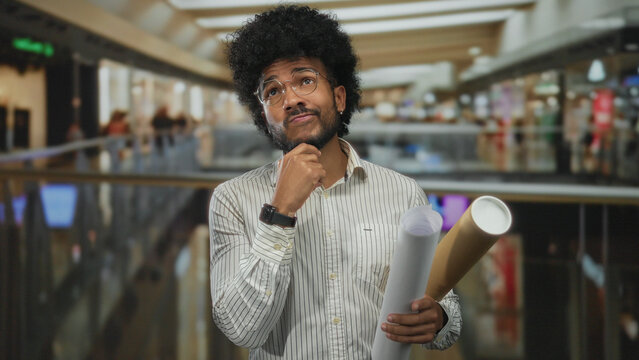 African american man holding blueprints in an indoor art gallery, appearing thoughtful and engaged.
