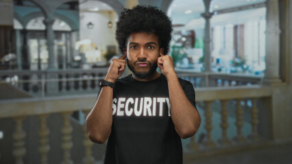 Security man standing indoors at a hotel, wearing a security uniform, covering his ears with both...
