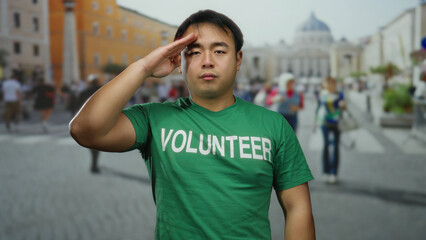 Asian man in green volunteer shirt saluting in st. peter's square, vatican city, with blurred background of people and historical architecture.