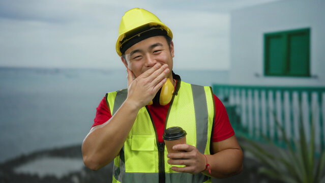 Man wearing safety vest and hard hat holding coffee on seaside promenade with building in background