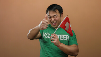 Young asian man holding swiss flag posing in front of isolated brown wall wearing volunteer shirt and expressing excitement with a thumbs-up gesture