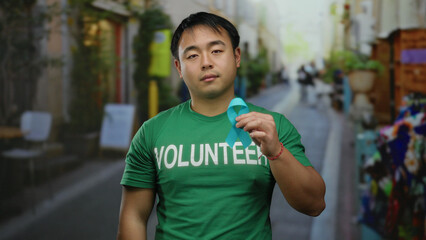 Young man wearing volunteer shirt holding blue ribbon on city street raising cancer awareness in...