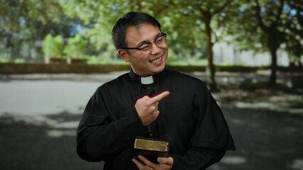 Asian priest smiling in park while holding bible, wearing black clerical attire under shade of...
