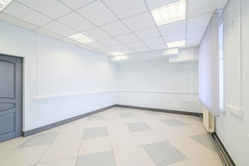 empty office room with white walls, a tiled floor, and a suspended ceiling with bright lights. A...