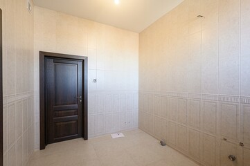empty room with beige tiled walls and floor, featuring a dark wood door and bright overhead lighting